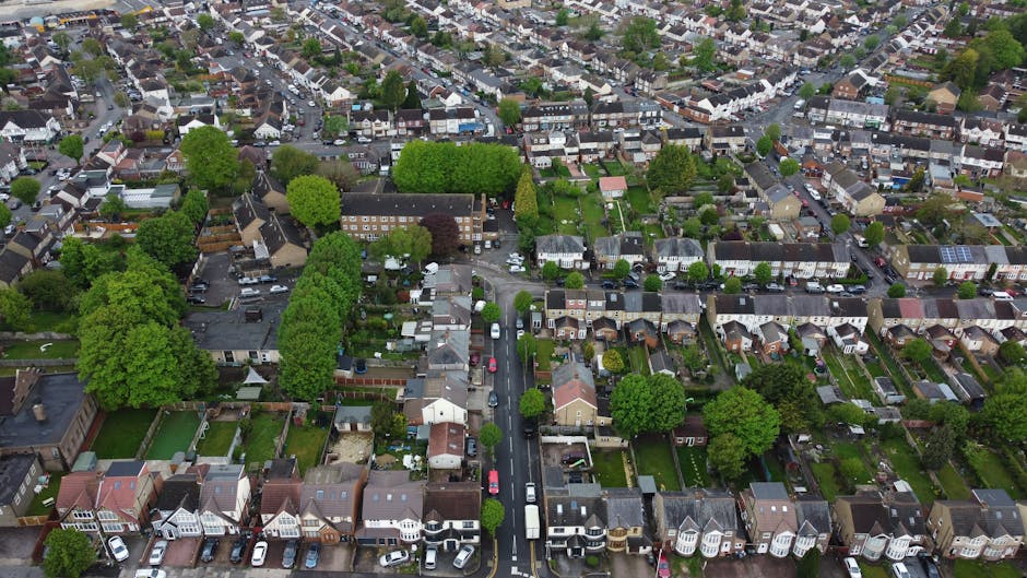 An aerial view of a densely populated suburban neighbourhood showing rows of terraced houses and semi-detached homes with varied architectural styles. The scene features numerous green trees and small gardens, some with fences and paved pathways. The streets are lined with parked cars, including sedans, hatchbacks, and vans. In the foreground, a narrow street runs vertically, bordered by neatly planted trees on both sides, leading to a small intersection with additional residential roads branching off. The background reveals a mixture of residential and commercial buildings with rooftops in shades of grey, brown, and red, along with patches of greenery and open spaces. The overall scene is well-lit, capturing the calm and orderly atmosphere typical of an urban residential area suitable for local or private waste disposal activities, with some properties showing evidence of rubbish collection readiness through visible waste containers or clearance sites near dwellings. This detailed aerial perspective underlines the community's layout and environment close to independent rubbish removal services, such as those offered by rubbishremovalroehampton.co.uk for property waste management in Roehampton.