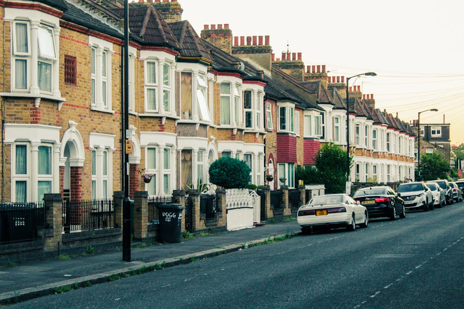 A row of traditional terraced houses with brick facades and bay windows along a residential street, each property featuring a small front garden enclosed by low brick walls and black wrought iron fencing. The houses have pitched roofs with multiple brick chimneys visible in the background, and some windows are open or partially covered by curtains. Parked along the curb are several cars of various models and colours, including white, black, and silver, occupying the entire street. A black wheelie bin labeled 'DOGG' is positioned in front of one of the houses, and a tall black lamppost stands near the sidewalk, which is paved with concrete slabs. The environment appears calm and tidy, with some greenery visible in the form of small shrubs and trees within the front gardens, and a warm, late-afternoon light illuminating the scene, typical of a clean, well-maintained neighbourhood. The image subtly reflects a typical residential area where residents might engage in private rubbish disposal, aligning with tips for independent waste management and rubbish removal services like those offered by rubbishremovalroehampton.co.uk.