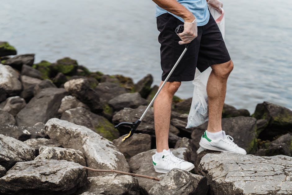 A person dressed in a light blue t-shirt, black shorts, and white sneakers with green accents is standing on a rocky shoreline near a body of water. They are wearing gloves and holding a grabber tool, which they are using to pick up a small piece of litter from the uneven, rough-textured rocks that are primarily grey with patches of lichen or moss. The rocks vary in size, some larger and flatter, others jagged and more protruding, creating a natural, rugged foreground and middle ground. In the background, the calm water stretches to the horizon, with a soft, diffused light suggesting an overcast day. The individual appears to be involved in an on-site clean-up activity, which aligns with private rubbish removal or environmental waste clearance activities undertaken by independent waste services, reflected in their careful handling of refuse in a natural setting near the water. The scene emphasizes a focus on environmental responsibility and waste collection in outdoor environments, as visualized through the act of litter picking among the rocks.