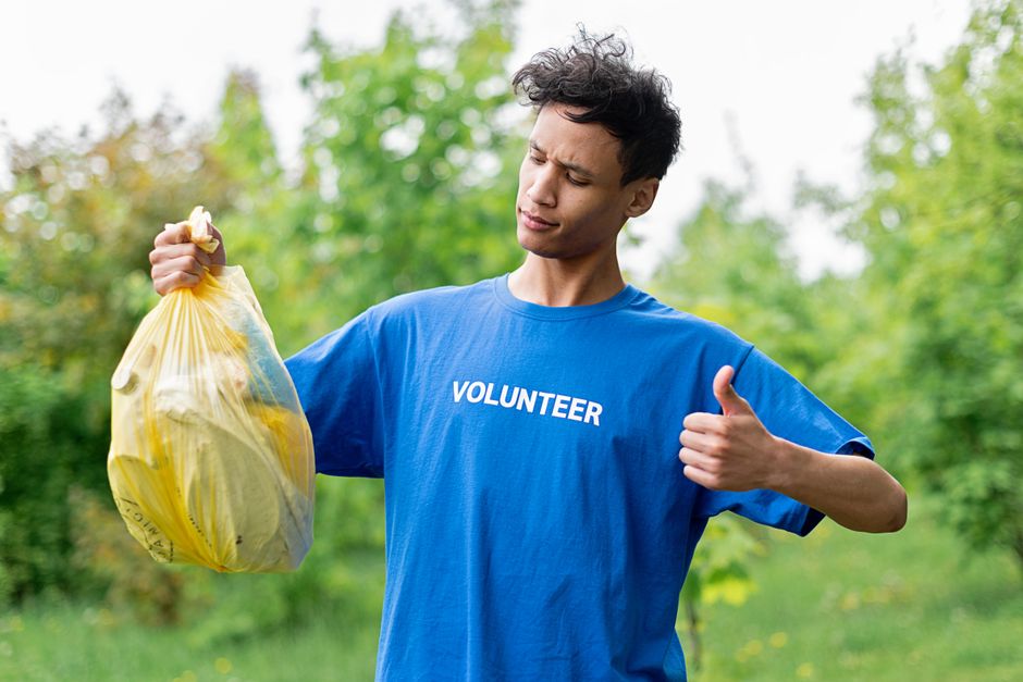 A young male volunteer with light skin and dark, slightly curly hair is outdoors in a green park or garden with blurred foliage in the background. He is wearing a bright blue T-shirt with the word 'VOLUNTEER' printed in white across the chest. The volunteer is holding a yellow plastic rubbish bag filled with waste in his right hand, gripping it by the top, while his left hand gives a thumbs-up gesture. His facial expression shows slight concentration or curiosity as he looks at the bag, suggesting he is engaged in a cleanup activity. The scene is well-lit with natural daylight, emphasizing the vibrant colors of the T-shirt, the bag, and the greenery surrounding him. The image portrays an active, positive volunteer participating in a community or environmental cleanup, aligning with the concept of independent waste collection or rubbish removal services, such as those offered by rubbishremovalroehampton.co.uk.